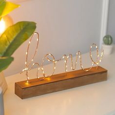 a wooden sign that says love is lit up with fairy lights and sits on a table next to a potted plant