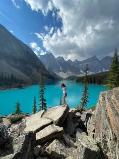 a woman standing on the edge of a cliff overlooking a lake with mountains in the background