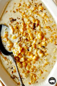 a white bowl filled with corn and cream on top of a wooden table next to a spoon