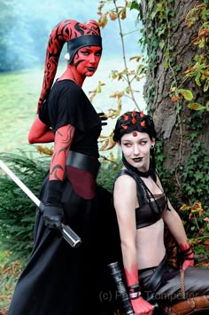 two women dressed in costumes standing next to each other near a tree and grass covered field