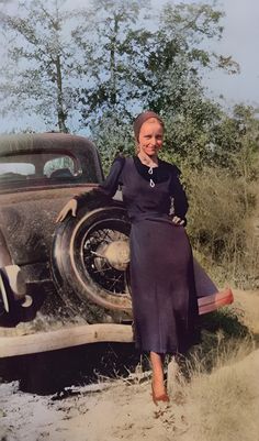 a woman standing next to an old car in the sand with trees and bushes behind her