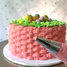 a cake being decorated with pink icing and green grass on top, while someone is using a sharp knife to cut it