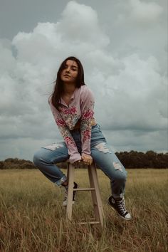 a young woman sitting on top of a wooden stool in a field with clouds above her