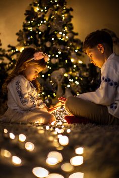 two children sitting on the floor in front of a christmas tree with lit candles around them