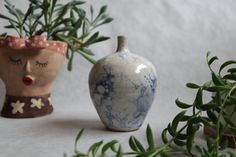 two ceramic vases sitting next to each other on a white surface with plants in the background