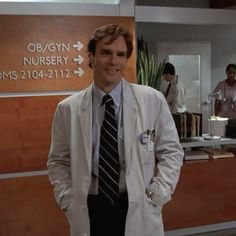 a man in a lab coat and tie standing next to a reception desk with people behind him