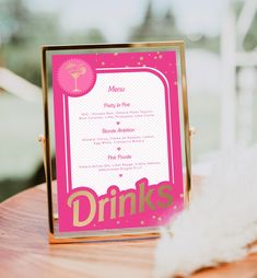 a pink and gold drink menu sitting on top of a wooden table next to a white feather