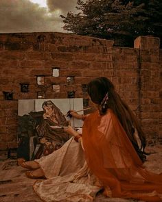 a woman sitting on the ground next to a brick wall with an easel in front of her