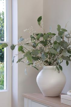 a white vase filled with flowers on top of a wooden table next to a window