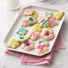 a white tray filled with colorful cookies next to a glass of milk on top of a table