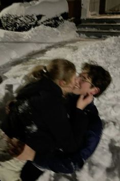 two people sitting in the snow with their faces close to each other and one person kissing