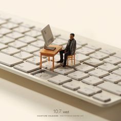 a miniature man sitting at a desk in front of a computer on top of a keyboard