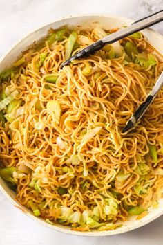 a bowl filled with noodles and vegetables on top of a white tablecloth next to two tongs