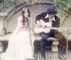 a man and woman sitting on a bench next to each other while playing guitar in front of a stone wall