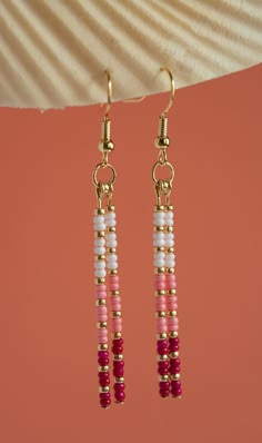 pink, white and gold beaded earrings hanging from a hook on a coral colored background