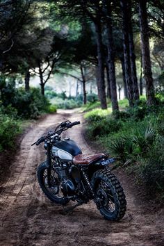 a motorcycle parked on the side of a dirt road near some trees and bushes,