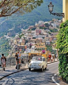 an old car is driving down the road in front of some buildings and people walking