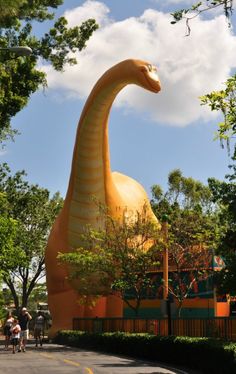 a large yellow dinosaur statue in the middle of a road with people walking by it