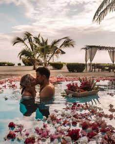 a man and woman kissing in a pool with flowers all over the water, surrounded by palm trees