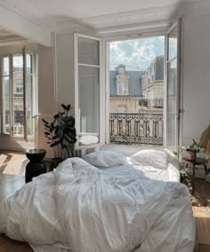 an unmade bed with white sheets and pillows in front of two large windows overlooking the city