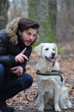 a man kneeling down next to a white dog on a leash and pointing at the camera