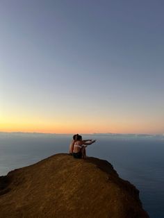 two people sitting on the edge of a cliff overlooking the ocean at sunset or sunrise