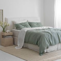 a bed with green sheets and pillows in a white room next to a plant on the floor