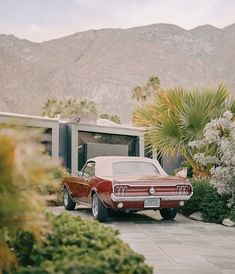 an old red car parked in front of a house with palm trees and mountains in the background