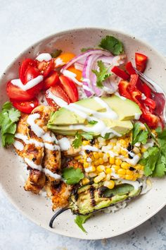 a white bowl filled with grilled chicken, vegetables and dressing on top of a table