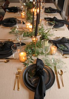 the table is set with black plates and silverware, greenery, candles and napkins