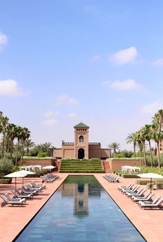 an empty pool with lounge chairs and umbrellas in front of a large building surrounded by palm trees