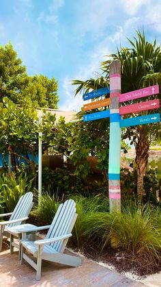 two white chairs sitting next to each other near a tree and grass covered area with palm trees in the background