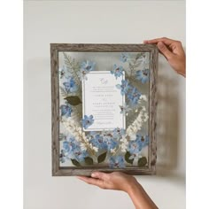 a person holding up a frame with blue flowers in it and a wedding card on the inside