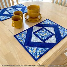 a wooden table topped with blue and yellow quilted placemats next to cups