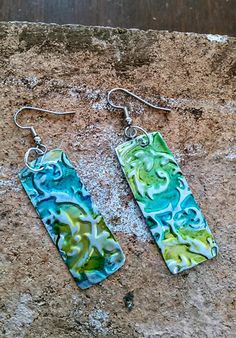 two rectangular earrings with green and blue swirls on them sitting on a stone surface