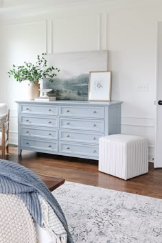 a blue dresser in a white bedroom with a plant on top and an ottoman next to it