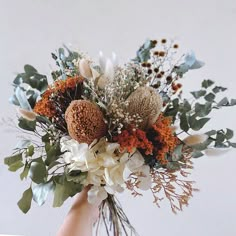 a hand holding a bouquet of flowers and greenery in front of a white wall