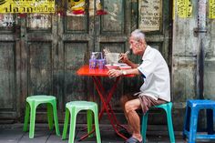 an old man sitting at a red table with three plastic chairs around him and eating