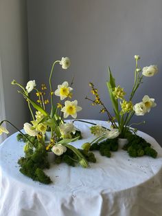 three vases with flowers and moss on a white table cloth covered round table in front of a window