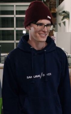 a young man wearing glasses and a beanie smiles at the camera while standing in front of a refrigerator