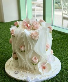 a white wedding cake with pink flowers on the top is sitting on green grass in front of a window