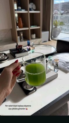 a woman holding a glass with green liquid in it on a counter top next to a window