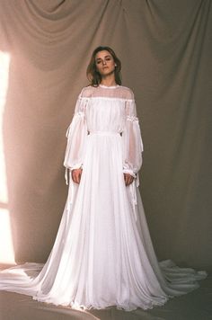 a woman standing in front of a curtain wearing a white dress with long sleeves and an open back