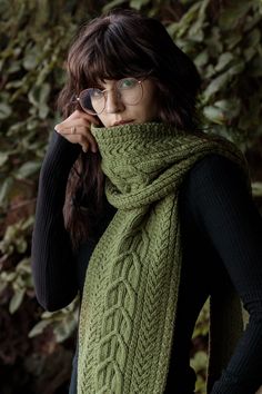 a woman wearing a green knitted scarf in front of a bush with leaves on it