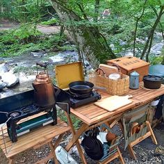 an outdoor table with pots and pans on it next to a stream in the woods