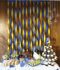 a table topped with cupcakes and cake next to a wall covered in blue and yellow ribbons