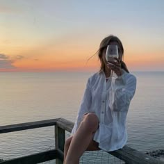 a woman sitting on a wooden railing drinking from a wine glass with the ocean in the background