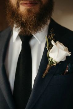 a bearded man wearing a suit and tie with a white rose pinned to his lapel