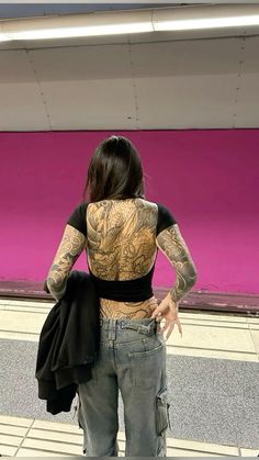 a woman with tattoos on her back standing in front of a pink wall and looking at the ground