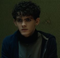 a young man with curly hair wearing a black jacket and grey sweater looking at the camera